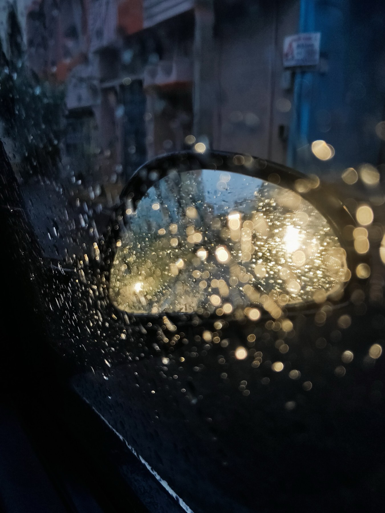 Rain beaded on a car window at night, a wing mirror lit by the city